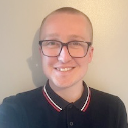 Customer Service Administrator Adam, a young man with a shaved head and glasses, smiling at the camera. He is wearing a dark polo shirt with a red and white trim and standing against a plain light-coloured wall.