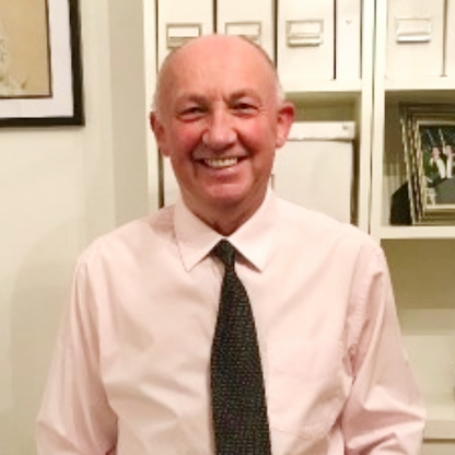 John Biggs, Trustee for BWBF, smiling while standing indoors in front of white shelving units. He is wearing a pale pink shirt with a dark patterned tie. Framed artwork and family photographs can be seen in the background.
