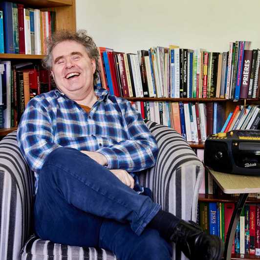 BWBF service user Dyfrig sits in a chair in a room with bookshelves. He is laughing and his Concerto 3 radio from BWBF is on a table next to him