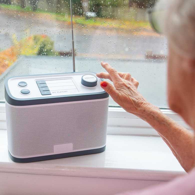 The Relish Radio sits on a windowsill as a user operates it. The radio is grey and white, with on/off button, four preset buttons and a volume control on top.