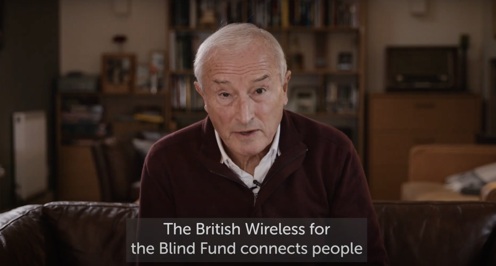 A photo of Jim Rosenthal sitting on a sofa as he talks to camera. A caption underneath reads 'The British Wireless for the Blind Fund connects people"