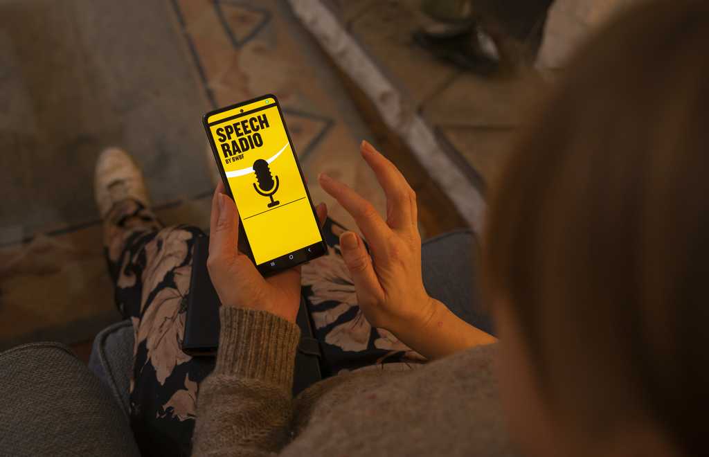 A person is browsing BWBF's Speech Radio app in a dimly lit room. The screen is lit up in yellow with the words 'Speech Radio' and an image of a microphone in black.