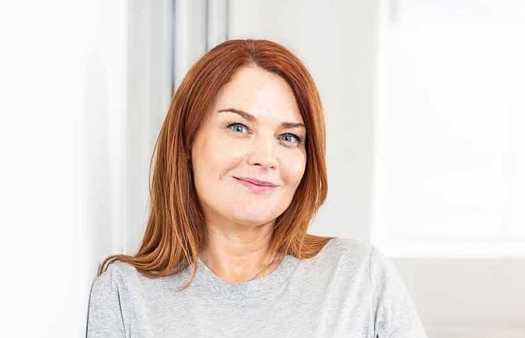 Sophie Jones, Chief Executive Officer at BWBF is smiling and leaning on a wall. She is wearing a pale grey top and her hair is dark red.