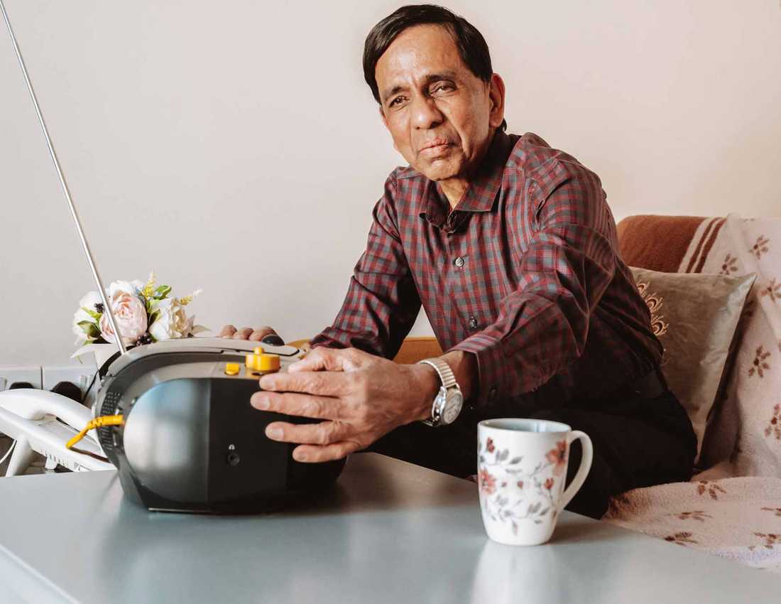 BWBF service user Krisha sits on a sofa. His hand is resting on his Concerto 3 radio from BWBF, which is on the table in front of him.