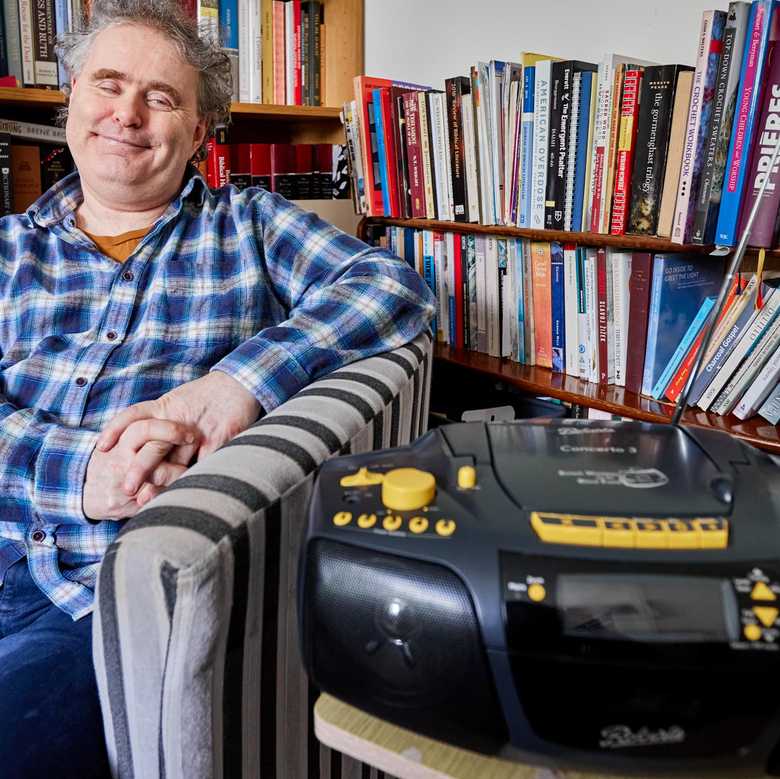 BWBF service user Dyfrig sits in a chair in a room with bookshelves. His Concerto 3 radio from BWBF is on a table next to him