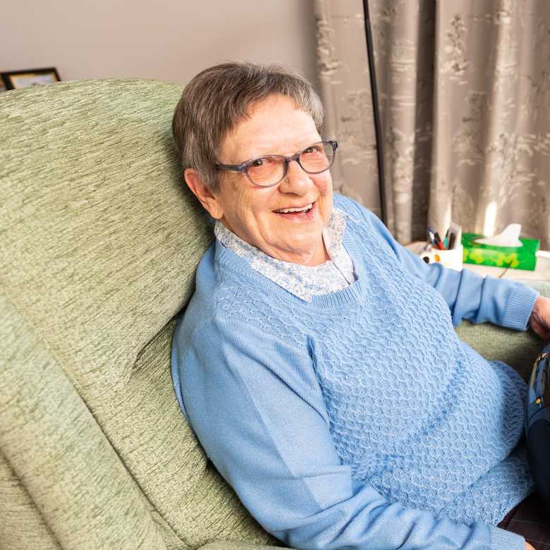Norma sits on an armchair. She has short grey hair and glasses and her Concerto 3 radio from BWBF is on her lap