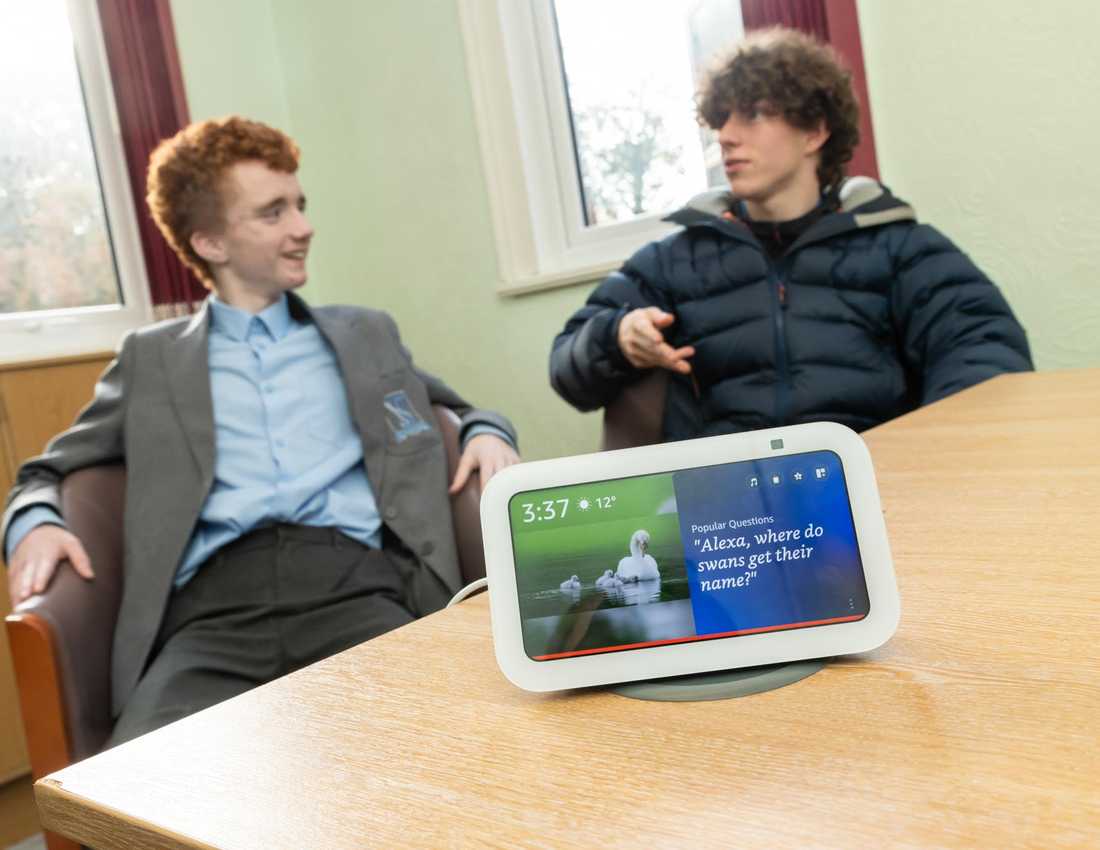 A white Alexa Show device with two teenage boys in the background, discussing something.