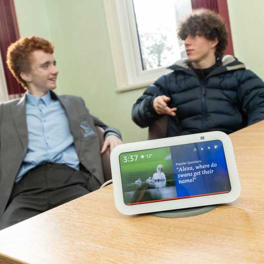 A white Alexa Show device with two teenage boys in the background, discussing something.