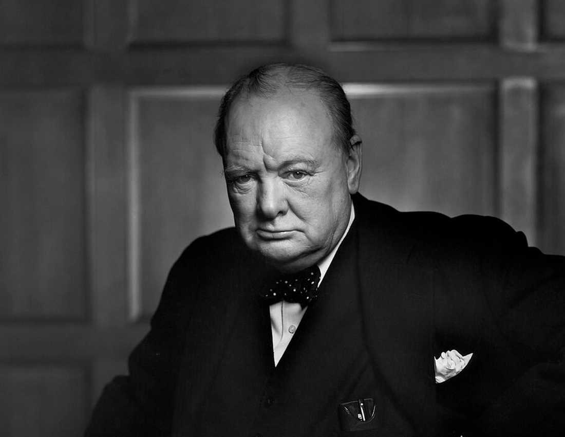 A black and white photo of Sir Winston Churchill sitting wearing a formal suit and bow tie. He has a cane in his hand.