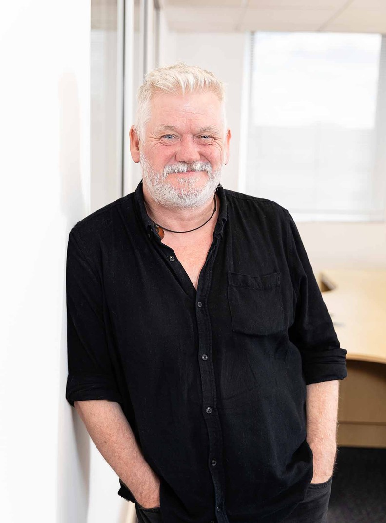 Steven Poole, Service Delivery Manager for BWBF, is leaning on a wall and smiling. He has wide grey hair and stubble is wearing a black shirt.