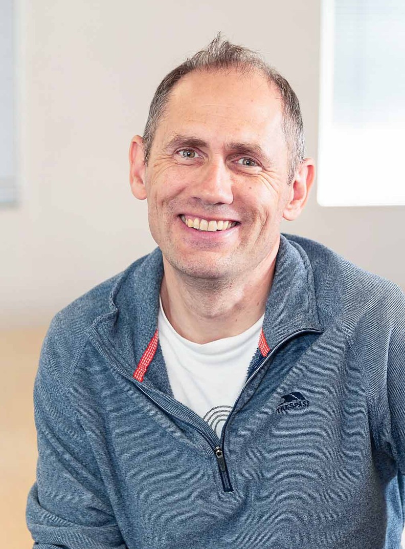 Simon Luget, Financial Controller for BWBF, sits on a table and is smiling. He has short grey hair and is wearing a bale blue casual top.