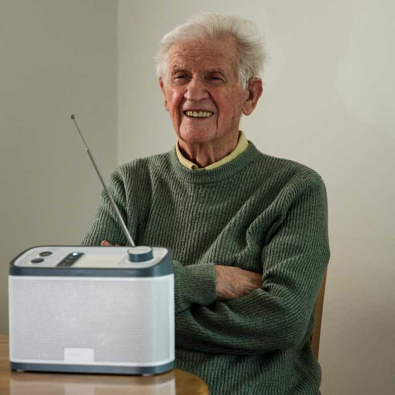 Thomas sits at a table smiling with his Relish radio from BWBF in front of him. His arms are folded.