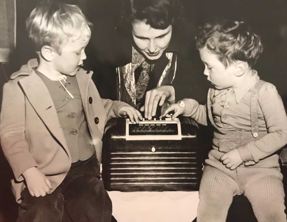 A black and white historical photo of a mother and her two sons. They are all pressing the buttons on the top of a large radio.