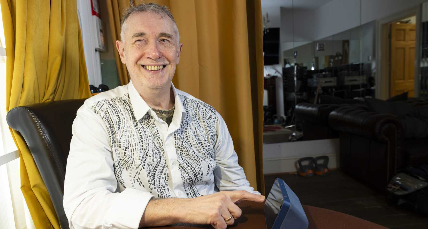 Paul Quigley, musician and performer. Paul lives with sight loss and is a beneficiary of BWBF. He is seated, smiling, as he engages with an Echo Show device on the table in front of him.