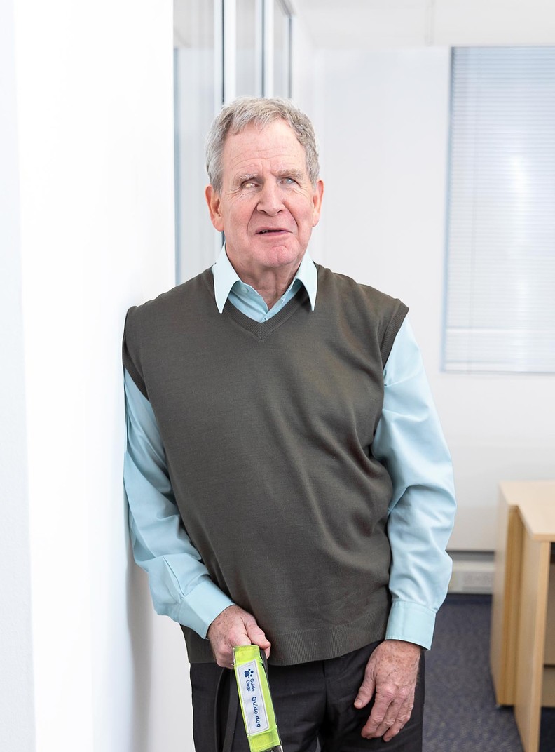 Mike Brace, Chair of BWBF, photographed in 2024. He is pictured leaning against a wall in an office and holding the lead for his guide dog.