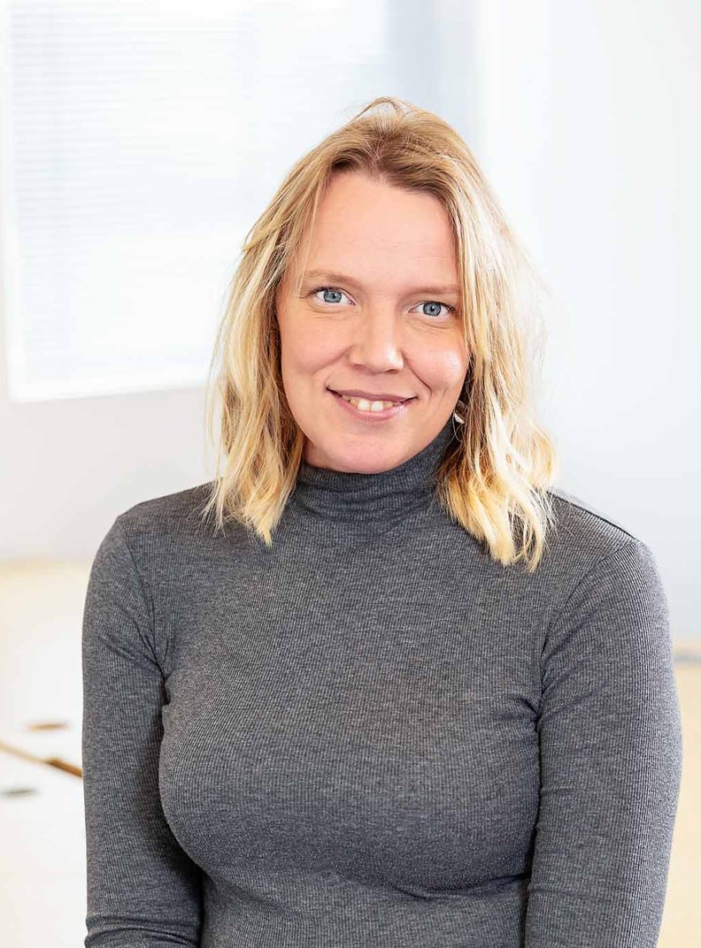 Jenny Cameron, Head of Communications, Marketing and Income for BWBF, sits on a table smiling to camera. She has blonde, wavy, shoulder-length hair and is wearing a grey top.