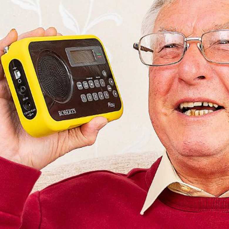 An older man with grey hair smiling as he holds a black Roberts Play radio with a yellow case. He is wearing a red jumper and glasses.