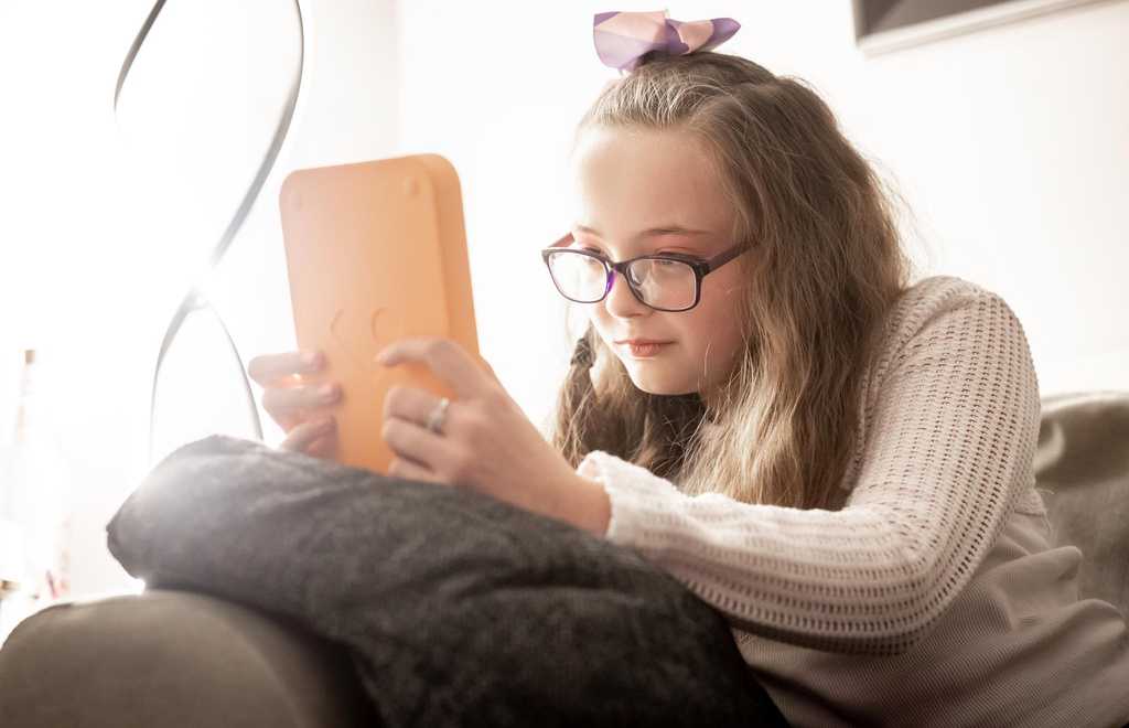 Annabel sits on her sofa reading her Voxblock audio book player. She wears glasses and has a bow in her hair.