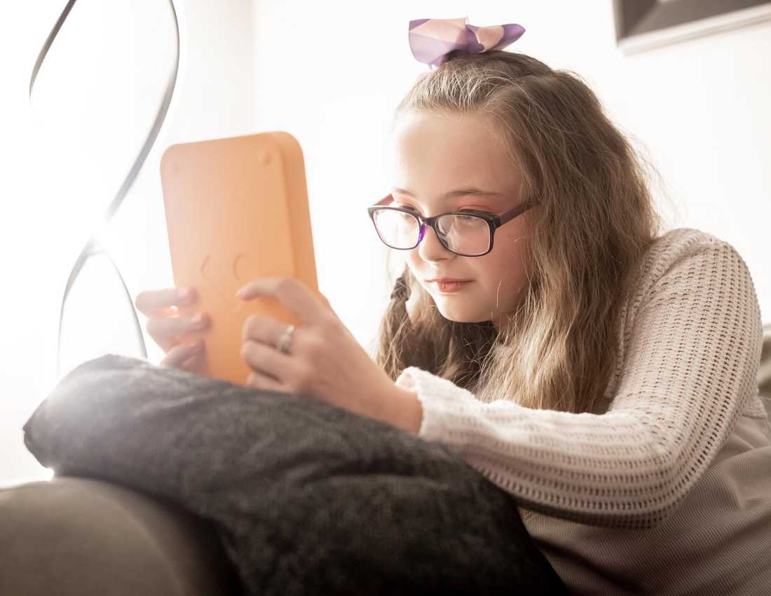 Annabel sits on her sofa reading her Voxblock audio book player. She wears glasses and has a bow in her hair.