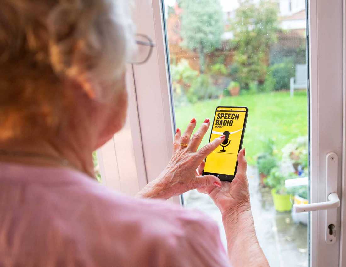 A woman stands in front of a window browsing BWBF's Speech Radio app. The app is black and yellow with an image of a microphone on it.