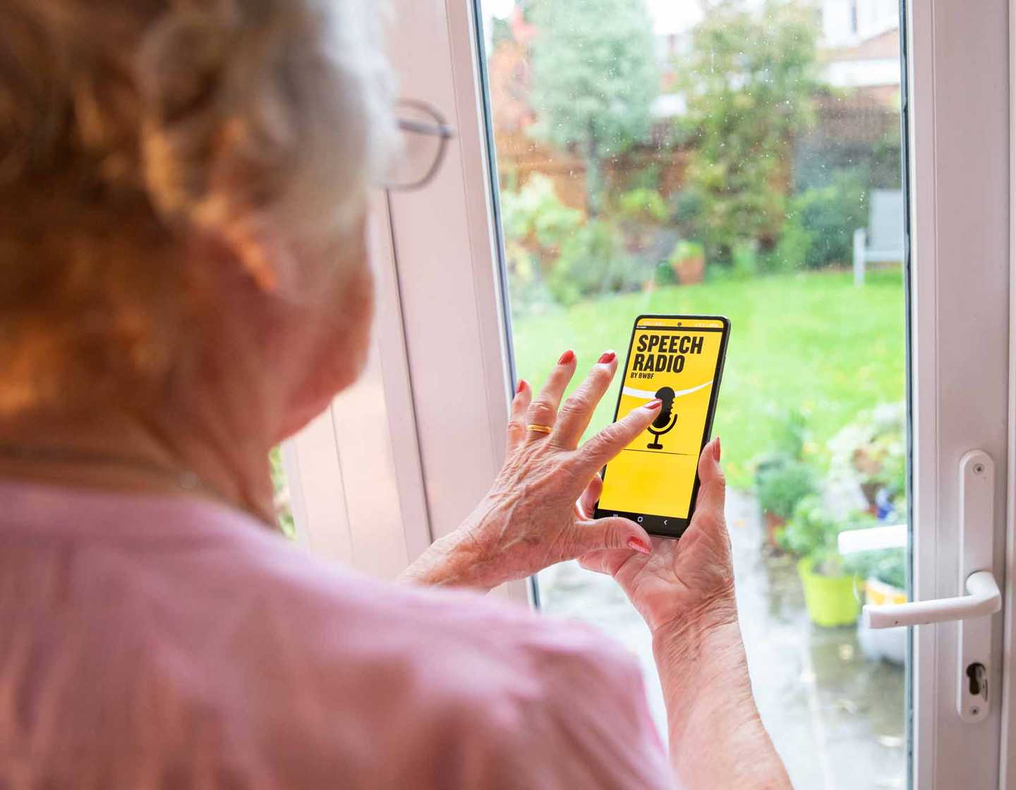 A woman stands in front of a window browsing BWBF's Speech Radio app. The app is black and yellow with an image of a microphone on it.