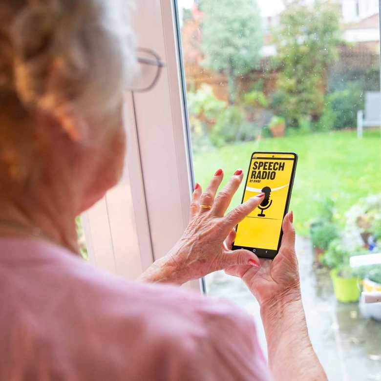 A woman stands in front of a window browsing BWBF's Speech Radio app. The app is black and yellow with an image of a microphone on it.