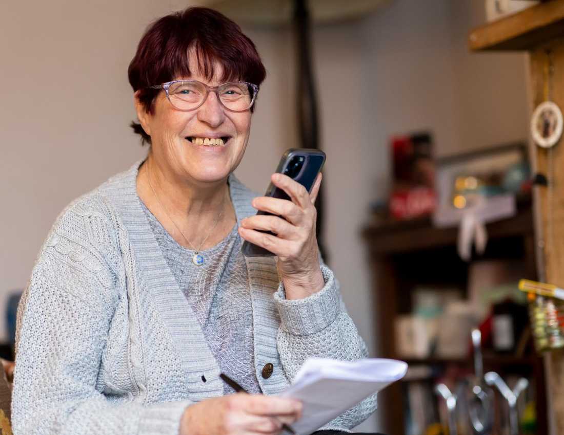 A woman stands in a kitchen smiling and as she talks on her phone which she is holding in front of her.