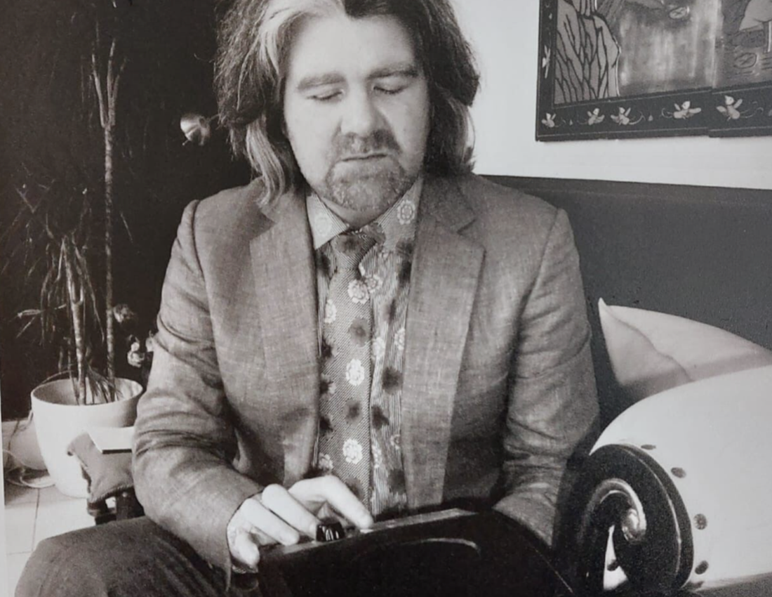 A black and white photo of a man with long grey hair sitting on a chair. He is pressing a button on a black Sonata audio device.
