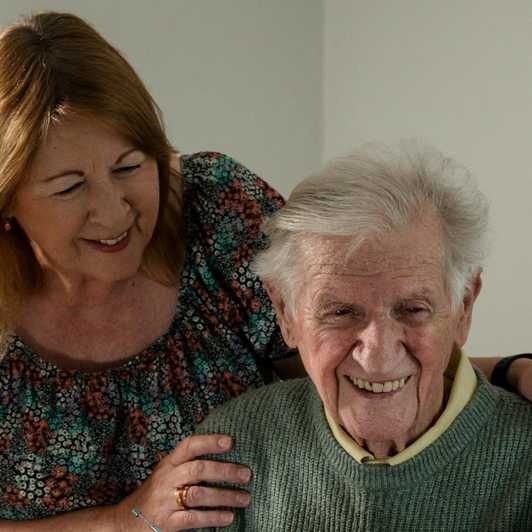 Thomas sits at a table smiling. His daughter Yvonne stands behind him looking at him and smiling, with her hands on his shoulder.