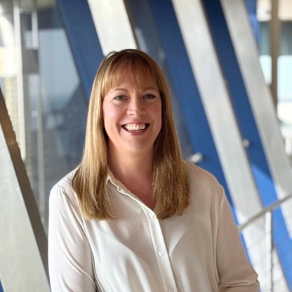 Alicia Coghlan, trustee for BWBF, smiling at the camera while standing indoors in a bright modern building with large blue and white structural beams in the background. She is wearing a white blouse and has shoulder-length light brown hair with a fringe.