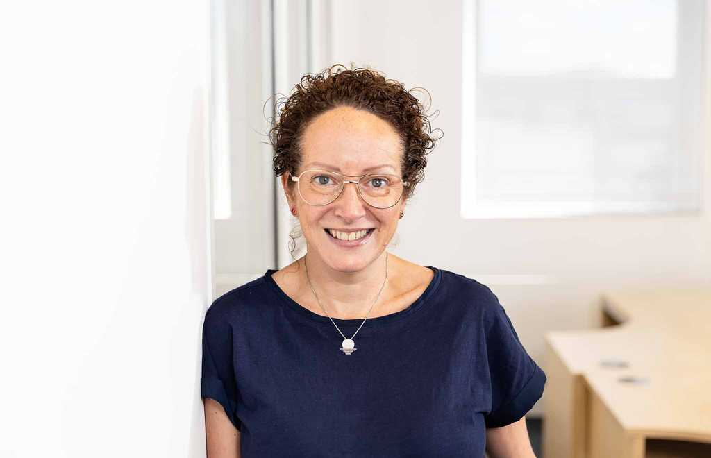 Julie Hocking, Individual Giving & Legacies Manager for BWBF, leans against a wall and is smiling. She wears glasses and her curly hair is tied up.